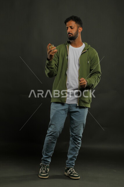 محاولة تحديد اسم المنتج, التسويق لمنتجات العطور, مفهوم التعطر والتطيب، عطورات خليجية سعودية, الاهتمام بالمظهر الخارجي, استخدام الزيوت العطرية الطبيعية, بورتريه لشاب عربي خليجي سعودي يرتدي الزي الكاجوال يحمل زجاجة العطر, صورة الجسم بالطول الكامل, خلفية سوداء