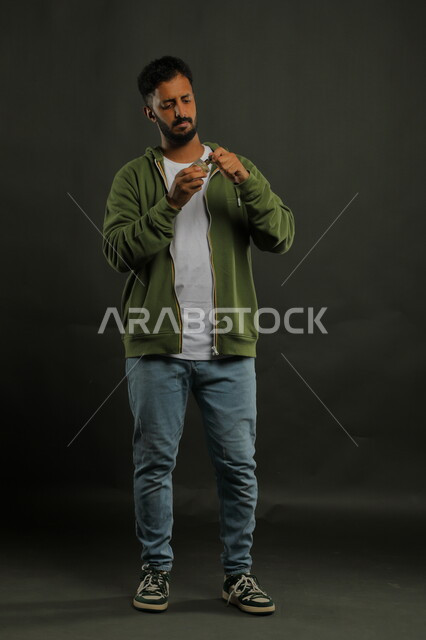 محاولة تحديد اسم المنتج, التسويق لمنتجات العطور, مفهوم التعطر والتطيب، عطورات خليجية سعودية, الاهتمام بالمظهر الخارجي, استخدام الزيوت العطرية الطبيعية, بورتريه لشاب عربي خليجي سعودي يرتدي الزي الكاجوال يحمل زجاجة العطر, صورة الجسم بالطول الكامل, خلفية سوداء