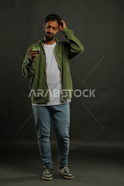 ألعاب تنمية الذكاء والمهارات العقليه, الاندماج والتركيز بنشاطات تقوية الذاكره، بورتريه لشاب وسيم عربي خليجي سعودي يرتدي الزي الكاجوال يمسك بيده المكعب السحري، الوقوف باستقامة بإيماءات التفكير والحيرة، صورة الجسم بالطول الكامل، خلفية سوداء
