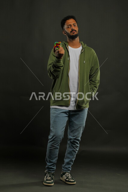 ممارسة الألعاب التعليمية الترفيهية، عرض تجربة اللعب بالروبيك كيوب، بورتريه لشاب وسيم عربي خليجي سعودي يرتدي الزي الكاجوال يمسك بيده مكعب مغناطيس السرعه، الوقوف باستقامة والنظر إلى الكاميرا، صورة الجسم بالطول الكامل، خلفية سوداء