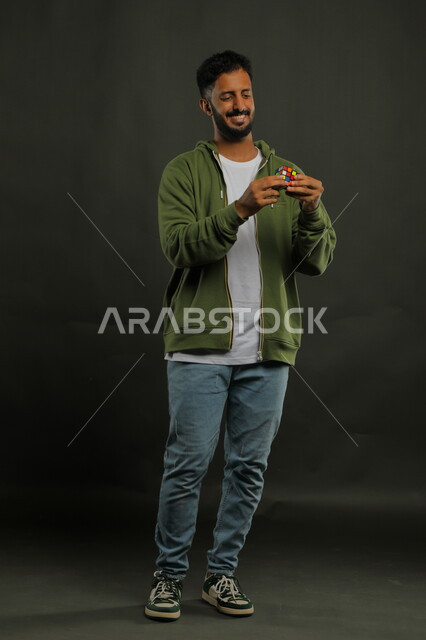 ممارسة الألعاب التعليمية الترفيهية، عرض تجربة اللعب بالروبيك كيوب، بورتريه لشاب وسيم عربي خليجي سعودي مبتسم يرتدي الزي الكاجوال يمسك بيده مكعب مغناطيس السرعه، الوقوف باستقامة وقضاء أوقات ممتعه في العطلة الصيفية، صورة الجسم بالطول الكامل، خلفية سوداء