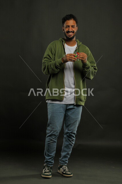 ممارسة الألعاب التعليمية الترفيهية، عرض تجربة اللعب بالروبيك كيوب، بورتريه لشاب وسيم عربي خليجي سعودي مبتسم يرتدي الزي الكاجوال يمسك بيده مكعب مغناطيس السرعه، الوقوف باستقامة وقضاء أوقات ممتعه في العطلة الصيفية، صورة الجسم بالطول الكامل، خلفية سوداء