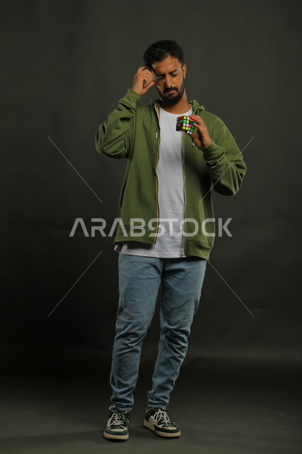 ألعاب تنمية الذكاء والمهارات العقليه, الاندماج والتركيز بنشاطات تقوية الذاكره، بورتريه لشاب وسيم عربي خليجي سعودي يرتدي الزي الكاجوال يمسك بيده المكعب السحري، الوقوف باستقامة بإيماءات التفكير والحيرة، صورة الجسم بالطول الكامل، خلفية سوداء