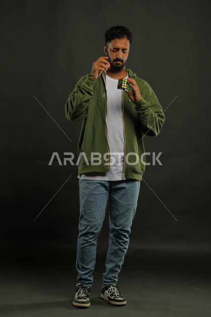 الاندماج والتركيز بنشاطات تقوية الذاكره, ألعاب تنمية الذكاء والمهارات العقليه، بورتريه لشاب وسيم عربي خليجي سعودي يرتدي الزي الكاجوال يمسك بيده المكعب السحري، الوقوف باستقامة بإيماءات التفكير والحيرة، صورة الجسم بالطول الكامل، خلفية سوداء