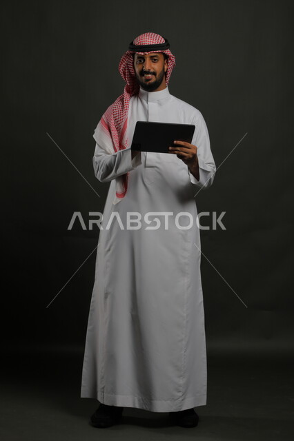 Using technology and modern techniques, browsing social networking sites, a portrait of a smiling Saudi Gulf Arab man wearing a shemagh and traditional thobe holding a tablet, chatting with family and friends, tablet applications and programs, full-length body portrait, gray background