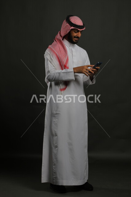 إيماءات السعادة لسماع الأخبار السارة, استخدام الهاتف المحمول، بورتريه لشاب عربي سعودي خليجي يرتدي الثوب والشماغ التقليدي مبتسم يغمض عينيه من شدة الفرح , التواصل مع الأهل والأصدقاء,  صورة الجسم بالكامل, خلفية رمادية.