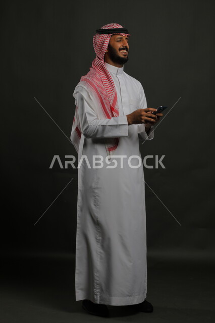 استخدام الهاتف المحمول، إيماءات السعادة لسماع الأخبار السارة, بورتريه لرجل عربي سعودي خليجي يرتدي الثوب والشماغ التقليدي مبتسم يغمض عينيه من شدة الفرح , التواصل مع الأهل والأصدقاء,  صورة الجسم بالكامل, خلفية رمادية.