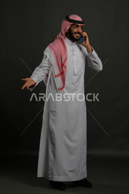 إيماءات الفرح بسماع خبر ما، إجراء مكالمات هاتفية باستخدام الهاتف المحمول, الشعور بالدهشة والسرور، بورتريه لشاب عربي خليجي سعودي مبتسم يرتدي الزي التقليدي يستخدم الجوال، صورة الجسم بالطول الكامل ,  خلفية رمادية .