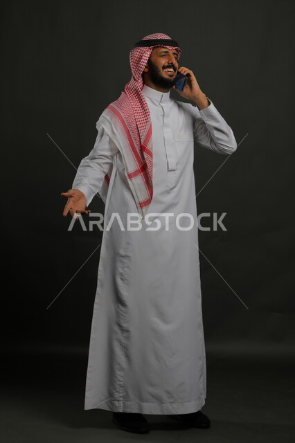 الشعور بالسعادة لسماع خبر ما، إجراء مكالمات هاتفية باستخدام الهاتف المحمول, التواصل مع الأهل والأصدقاء، بورتريه لشاب عربي خليجي سعودي مبتسم يرتدي الزي التقليدي يغمض عينيه من شده الفرح يستخدم الجوال، صورة الجسم بالطول الكامل ,  خلفية رمادية .