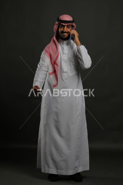 استخدام الهاتف المحمول لإجراء المكالمات الهاتفية, الشعور بالحماس والسرور, بورتريه لرجل عربي سعودي خليجي مبتسم يرتدي الثوب والشماغ التقليدي ينظر إلى الكاميرا يرفع يده بإيماءات الاندهاش والذهول, صورة الجسم بالكامل, خلفية رمادية.