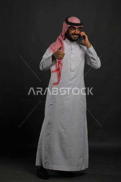 الشعور بالرضا والقبول, بورتريه لشاب عربي سعودي خليجي يرتدي الثوب والشماغ التقليدي مبتسم يستخدم الهاتف المحمول يرفع إصبعه نحو الأعلى بعلامة الجودة, تحقيق أهداف العمل, صورة الجسم بالكامل, خلفية رمادية.
