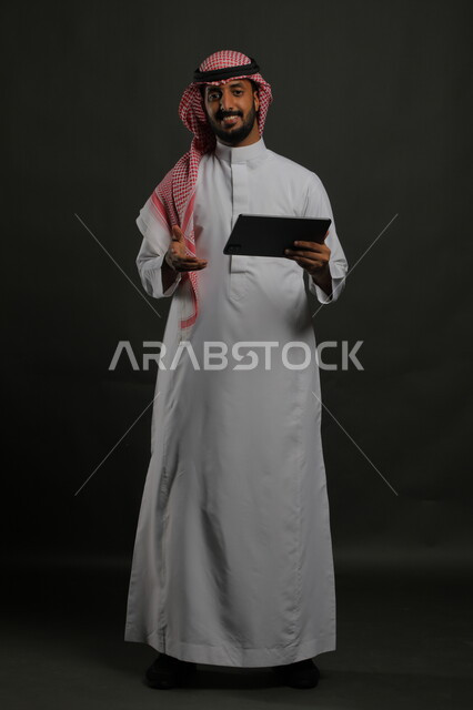 استخدام التكنولوجيا والتقنيات الحديثة في الشرح والتعليم, بورتريه لرجل عربي خليجي سعودي يرتدي الشماغ والثوب التقليدي يقدم البرزينتيشن في اجتماع العمل, استعمال جهاز التابليت في العرض التقديمي, صورة لكامل الجسم, خلفية سوداء.