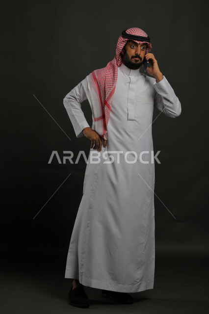 إيماءات الدهشة و الذهول، الصدمة والمفاجأة بسماع خبر ما، بورتريه لرجل عربي سعودي خليجي يرتدي الزي التقليدي والشماغ يضع يده على خصره يقوم بإجراء مكالمة صوتية عبر الهاتف المحمول، صورة الجسم بالطول الكامل، خلفية رمادية.