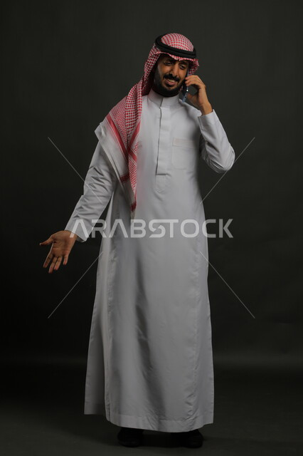 الاستفسار والسؤال عن أمر ما, بورتريه لرجل عربي سعودي خليجي مبتسم يرتدي الثوب والشماغ التقليدي يقف يستخدم الهاتف المحمول, التواصل مع الأهل والأصدقاء للاطمئنان, إجراء مكالمات صوتية, صورة الجسم بالكامل, خلفية رمادية.