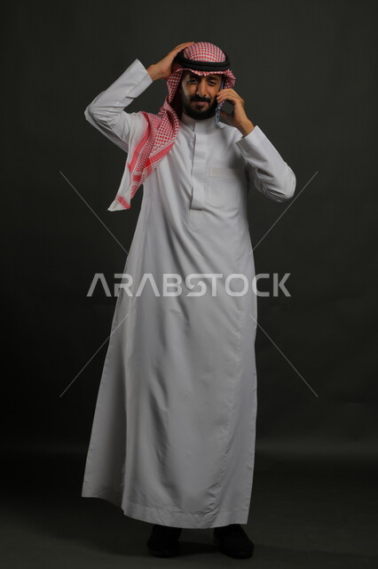 Gestures of thinking and contemplation to solve problems, portrait of a smiling Saudi Gulf Arab young man wearing a traditional thobe and shemagh, placing his hand on his head, using a mobile phone, discussing and arguing about something, making voice calls, full body image, gray background