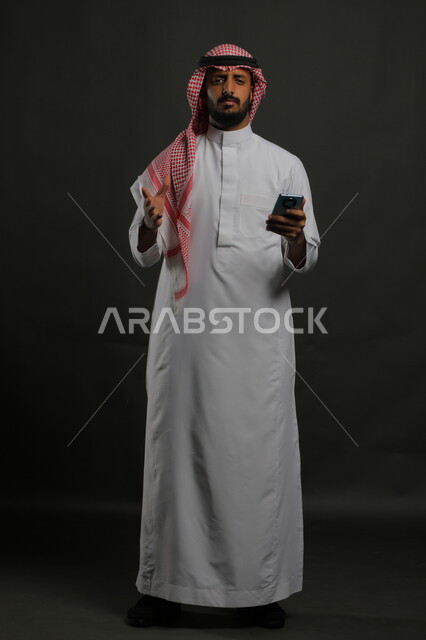Grimacing, angry, and frowning to see something, feeling upset to hear bad news, portrait of a Saudi Gulf Arab young man wearing a shemagh and traditional dress, with his hand on his waist, using a mobile phone, full-length body portrait, gray background