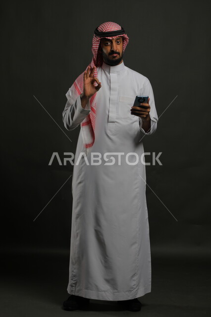 الإعجاب بالتقنيات الحديثة, بورتريه لشاب عربي سعودي خليجي يرتدي الثوب والشماغ التقليدي مبتسم يستخدم الهاتف المحمول ينظر إلى الكاميرا, إيماءات جودة الأجهزة الحديثة وتطبيقاتها, صورة الجسم بالكامل, خلفية رمادية.