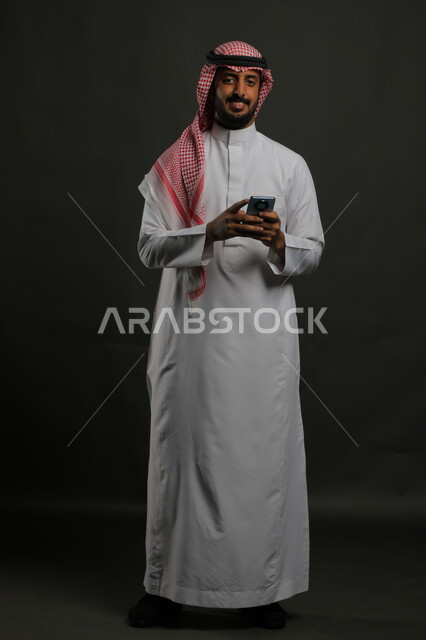 Gestures of happiness to hear good news, using a mobile phone, portrait of a Saudi Gulf Arab young man wearing the traditional thobe and shemagh, smiling, closing his eyes with joy, communicating with family and friends, full body portrait, gray background