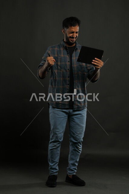 مشاهدة مباريات كرة القدم عبر الجهاز اللوحي، التواصل مع الأهل والأصدقاء وسماع الأخبار السارة, بورتريه لشاب عربي سعودي خليجي يرتدي الزي الكاجوال يستخدم التابلت يرفع قبضته بإيماءات الحماس والسرور بفوز الفريق المفضل, صورة الجسم بالكامل, خلفية رمادية.