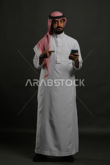 إيماءات جودة الأجهزة الحديثة وتطبيقاتها, الأداء الفعال للتقنيات المتطورة, بورتريه لشاب عربي سعودي خليجي يرتدي الثوب والشماغ التقليدي ينظر إلى الكاميرا يشير بإصبعه إلى الهاتف المحمول, صورة الجسم بالكامل, خلفية رمادية.