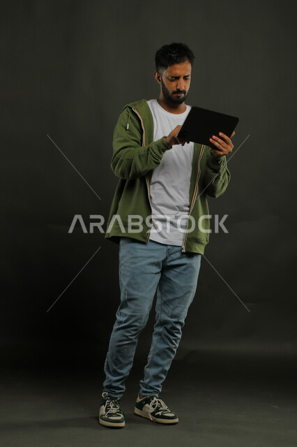 تلقي أخبار مزعجة من العمل، الشعور بالانزعاج لمعرفة الأخبار السيئة, إيماءات الغضب والعبوس, بورتريه لشاب عربي سعودي خليجي يرتدي اللباس الكاجوال يستخدم جهاز التابليت, دردشات ومحادثات عبر الانترنت, صورة الجسم بالكامل, خلفية سوداء