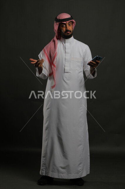 أمر مجهول ومبهم,  إجراء المكالمات الصوتية عبر الهاتف المحمول، بورتريه لشاب عربي سعودي خليجي يرتدي الثوب والشماغ التقليدي ينظر إلى الكاميرا يرفع كفه نحو الأعلى بإيماءات الجهل وعدم المعرفة, صورة الجسم بالكامل, خلفية رمادية.