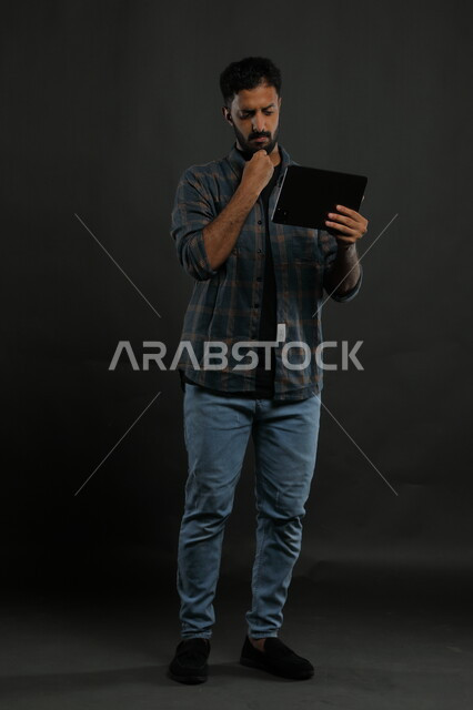 احتراف اوظائف التفنية وتكنولوجيا المعلومات، مشاهدة أخبار مزعجة على وسائل التواصل الاجتماعي، بورتريه لشاب عربي سعودي خليجي يرتدي اللباس الكاجوال يستخدم جهاز التابليت بإيماءات العبوس والانزعاج, صورة الجسم بالكامل, خلفية سوداء