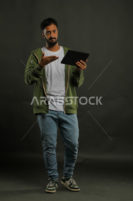 الأداء الفعال للتكنولوجيا والتقنيات المتطورة, بورتريه لشاب عربي سعودي خليجي يرتدي الزي الكاجوال ينظر إلى الكاميرا يشير بيده إلى الجهاز اللوحي, جودة الأجهزة الحديثة وتطبيقاتها, صورة الجسم بالكامل, خلفية سوداء