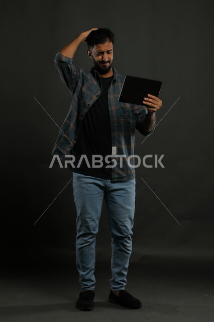 إنجاز مهام العمل عن بعد، تعابير الانزعاج بتلقي الأخبار السيئة، بورتريه لرجل عربي خليجي سعودي يرتدي الزي الكاجوال يحمل بيده جهاز التابلت ويضع يده على رأسه بإيماءات الغضب والعبوس، صورة الجسم بالكامل, خلفية سوداء