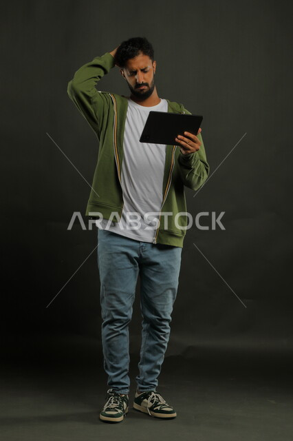 إنجاز مهام العمل عن بعد، تعابير الانزعاج بتلقي الأخبار السيئة، بورتريه لرجل عربي خليجي سعودي يرتدي الزي الكاجوال يحمل بيده جهاز التابلت ويضع يده على رأسه بإيماءات الغضب والعبوس، صورة الجسم بالكامل, خلفية سوداء