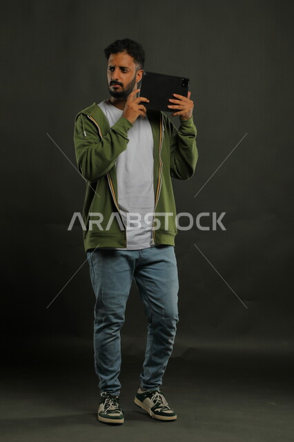 محاولة سماع التسجيلات والمقاطع الصوتية، اكتشاف مشاكل الصوت في الجهاز اللوحي، بورتريه لرجل عربي خليجي سعودي يرتدي الزي الكاجوال يحمل بيده جهاز التابلت بإيماءات العبوس والانزعاج لتصليح الأعطال التقنية، صورة الجسم بالكامل, خلفية سوداء