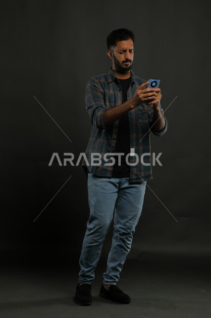 Following social media and websites, feeling disturbed when watching bad news, portrait of a Saudi Gulf Arab young man wearing a casual outfit using a mobile phone, gestures of anger, frowning, and anxiety, full-length body image, black background