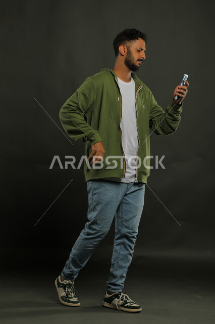 Following social media and websites, feeling disturbed when watching bad news, portrait of a Saudi Gulf Arab young man wearing a casual outfit using a mobile phone, gestures of anger, frowning, and anxiety, full-length body image, black background