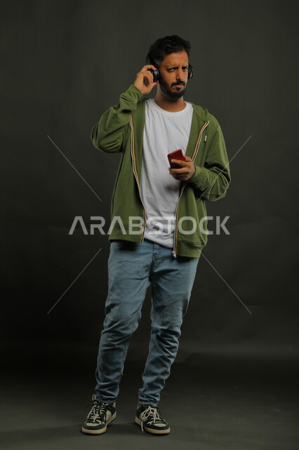 Diving into the world of music, gestures of bewilderment and astonishment, watching series and movies using a mobile phone, a portrait of a Saudi Gulf Arab young man wearing casual summer clothes pointing with his finger at his mobile phone, full-length body portrait, gray background