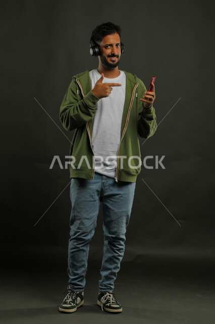 Gestures for enjoying music, watching series and movies using a mobile phone, portrait of a Saudi Gulf Arab young man wearing casual summer clothes pointing with his finger at his mobile phone, full-length body portrait, gray background