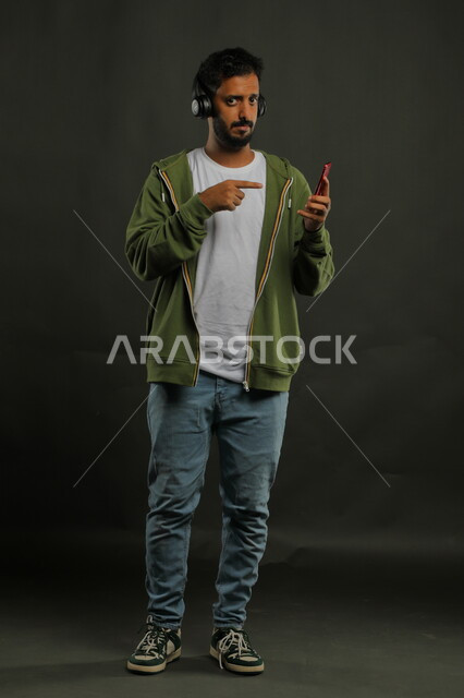 مشاهدة المسلسلات والأفلام باستخدام الجوال، ايماءات الحزن والكآبة،  بورتريه لشاب عربي سعودي خليجي يرتدي ملابس كاجوال  يشير باصبعه الى هاتفه المحمول، صورة الجسم بالطول الكامل، خلفية رمادي
