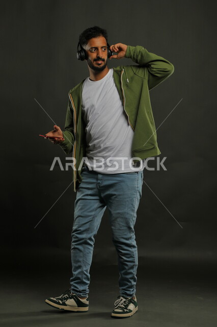  استخدام سماعات الهيدفون لقضاء وقت ممتع، ايماءات النظر والتركيز بشيء ما، بورتريه لشاب عربي سعودي خليجي يرتدي ملابس كاجوال يضع سماعات الرأس ويمسك بيده الهاتف المحمول، صورة الجسم بالكامل، خلفية رمادي
