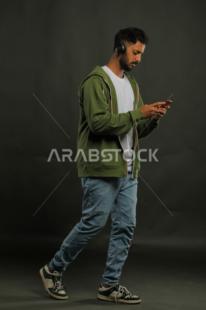 استخدام  تطبيقات الاغاني والتسجيلات الصوتية في الجوال، الدردشة والتحدث مع الأصدقاء عبر البث المباشر، بورتريه لشاب عربي سعودي خليجي يرتدي ملابس كاجوال يضع سماعات الهيدفون، صورة الجسم بالكامل، خلفية رمادي