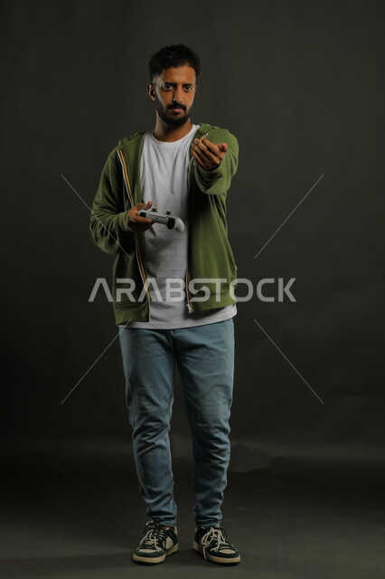 تجربة ترفيهية ممتعة ومثيرة في الألعاب الالكترونية ، الابتعاد عن الضغوط اليومية والاستمتاع بالوقت، بورتريه لشاب عربي سعودي خليجي يرتدي الزي الكاجوال يمسك يد التحكم بايماءات الغضب، العالم الرقمي الترفيهي، صورة الجسم بالطول الكامل، خلفية رمادية