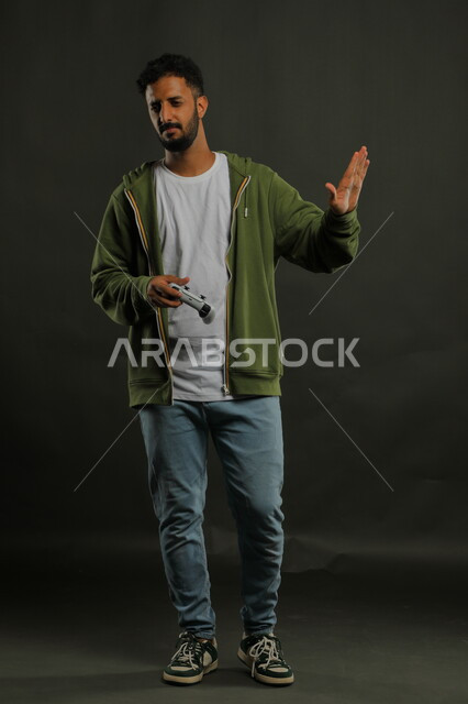 ايماءات طلب التوقف والابتعاد، قضاء وقت الفراغ بالألعاب الالكترونيه،  بورتريه لشاب عربي سعودي خليجي يرتدي الزي الكاجوال يمسك قبضة التحكم ، المتعة و التسلية بالفيديو جيمز،  صورة الجسم بالطول الكامل، خلفية رمادية