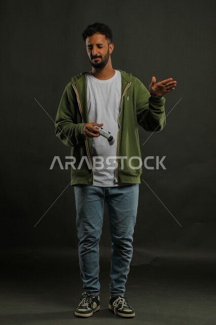 ايماءات الاستهزاء والسخرية، الابتعاد عن الضغوط اليومية وقضاء الوقت بالألعاب الالكترونيه،  بورتريه لشاب عربي سعودي خليجي يرتدي الزي الكاجوال يمسك قبضة التحكم، المتعة و التسلية بالفيديو جيمز،  صورة الجسم بالطول الكامل، خلفية رمادية