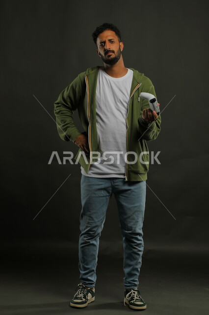 عدم تقبل الخسارة،  المتعة والتسلية  بالفيديو جيمز، الالعاب الالكترونيه الترفيهية والممتعة، بورتريه لرجل عربي سعودي خليجي يرتدي الزي الكاجوال يمسك يد التحكم ويضع يده على خصره بغضب،  صورة الجسم بالطول الكامل، خلفية رمادية