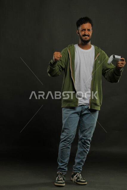 الالعاب الالكترونيه الممتعة، ايماءات الخسارة وخيبة الأمل، بورتريه لشاب عربي سعودي خليجي يرتدي الزي الكاجوال يمسك يد التحكم ويرفع قبضته للأعلى بغضب، المتعة والتسلية  بالفيديو جيمز،  صورة الجسم بالطول الكامل، خلفية رمادية