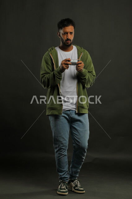 Gestures of integration and interaction with the video game, spending a good time with recreational electronic games, a portrait of a young Saudi Arabian Gulf man wearing a casual outfit holding the controller and looking with concentration and attention, full-length body portrait, gray background