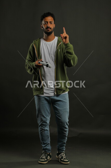 قضاء وقت ممتع  بالألعاب الالكترونية الترفيهية، بورتريه لشاب عربي سعودي خليجي يرتدي الزي الكاجوال يمسك يد التحكم ويشير الى الأعلى، إيماءات الإندماج والتفاعل مع الجيم، صورة الجسم بالطول الكامل، خلفية رمادية