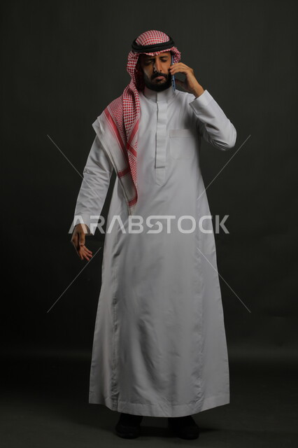 إيماءات الحزن بسماع الأخبار السيئة, الشعور بالإحباط واليأس, بورتريه لرجل عربي سعودي خليجي يرتدي الثوب والشماغ التقليدي يغمض عينيه يستخدم الهاتف المحمول, صورة الجسم بالكامل, خلفية رمادية.