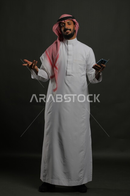 الاستفهام والاستفسار عن أمر ما, بورتريه لرجل عربي سعودي خليجي مبتسم يرتدي الثوب والشماغ التقليدي يقف يستخدم الهاتف المحمول, التواصل مع الأهل والأصدقاء للاطمئنان, إجراء مكالمات صوتية, صورة الجسم بالكامل, خلفية رمادية.