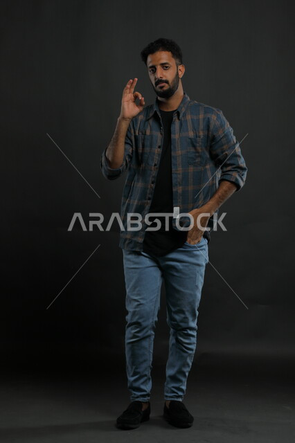 الشعور بالإعجاب والرضا والسعاده، النظر إلى الكاميرا بايماءات القبول والموافقه, بورتريه لشاب وسيم عربي خليجي سعودي مبتسم يرتدي الزي الكاجوال ويرفع أصبع الابهام بعلامة الجودة ،صورة الجسم بالطول الكامل، خلفية سوداء