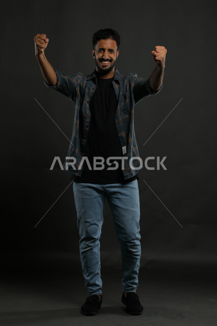 الشغف في تشجيع فريق كرة القدم، ايماءات الفوز والانتصار وتحقيق الأهداف ،حركات تدل على الكسب والربح وانجاز المهام ،بورتريه لشاب عربي خليجي سعودي يرتدي الزي الكاجوال يرفع يديه بتفاعل وحماس ، صورة الجسم بالطول الكامل، خلفية سوداء