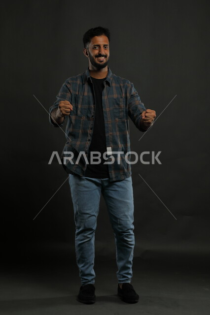 ايماءات الفوز والانتصار وتحقيق الأهداف ،حركات تدل على الكسب والربح وانجاز المهام ،بورتريه لشاب عربي خليجي سعودي يرتدي الزي الكاجوال يرفع يديه بتفاعل وحماس ،الاندماج بتشجيع فريق كرة القدم، صورة الجسم بالطول الكامل، خلفية سوداء
